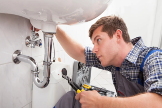 Un plombier professionnel en uniforme de travail, portant une ceinture à outils et travaillant sur des tuyaux sous un évier. Il utilise une clé pour resserrer un raccord, entouré de divers outils de plomberie comme des pinces et un ruban d'étanchéité.